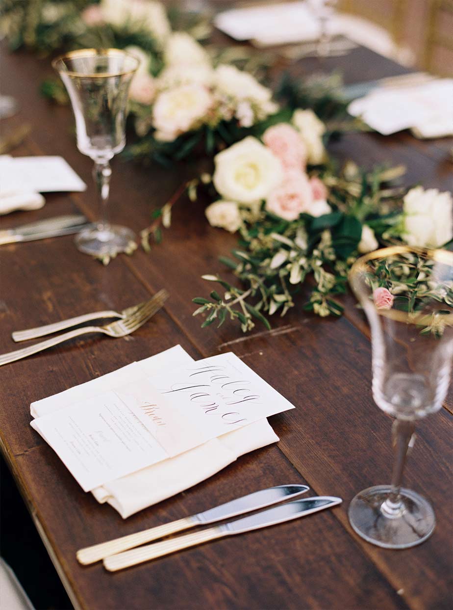 Wooden reception head table wedding place setting with gold rimmed glasses and floral garland