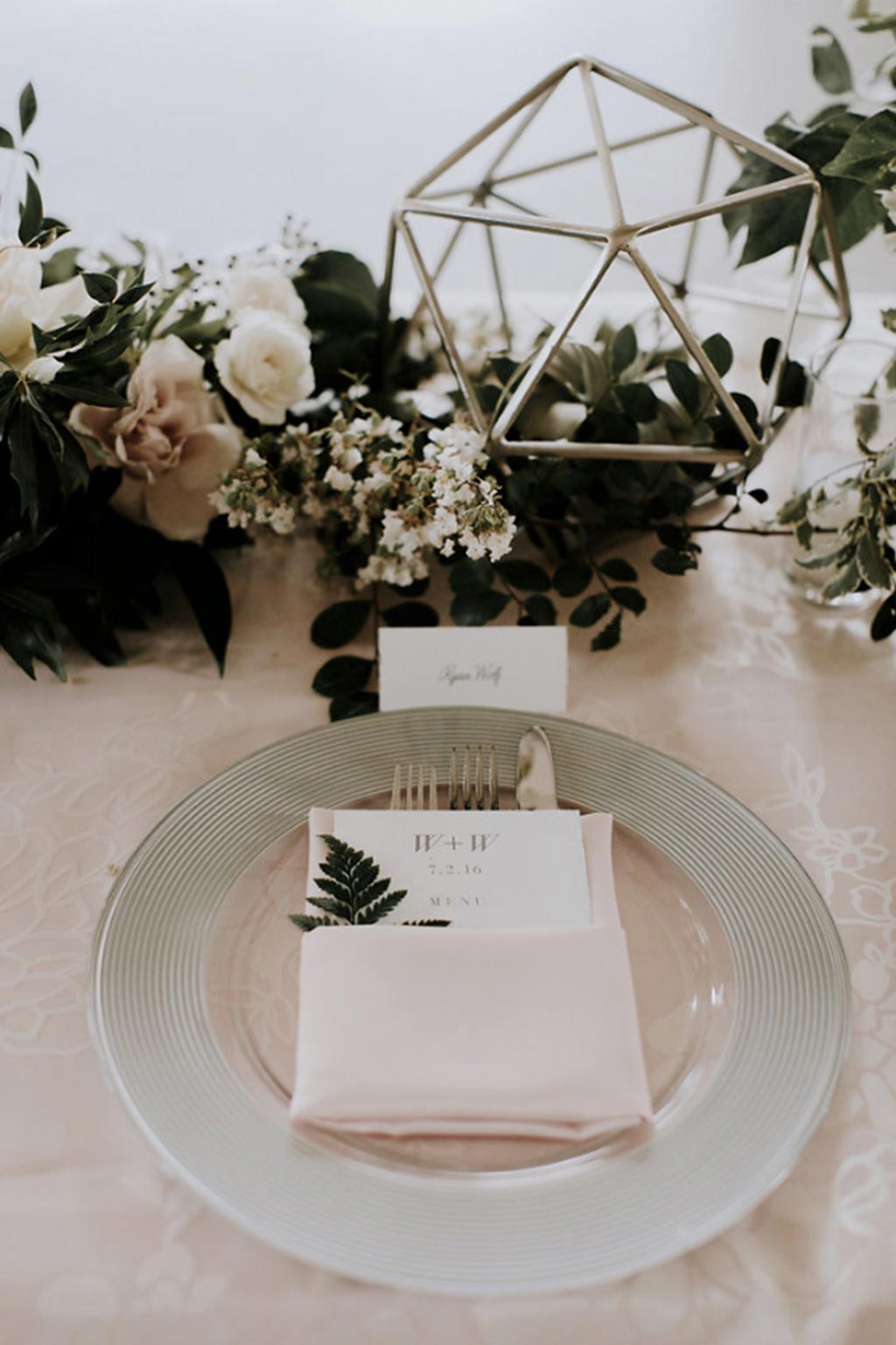 Faceted terrarium with green and white organic garland wedding centerpiece and silver and glass, pink napkin placesetting with menu and fern leaf